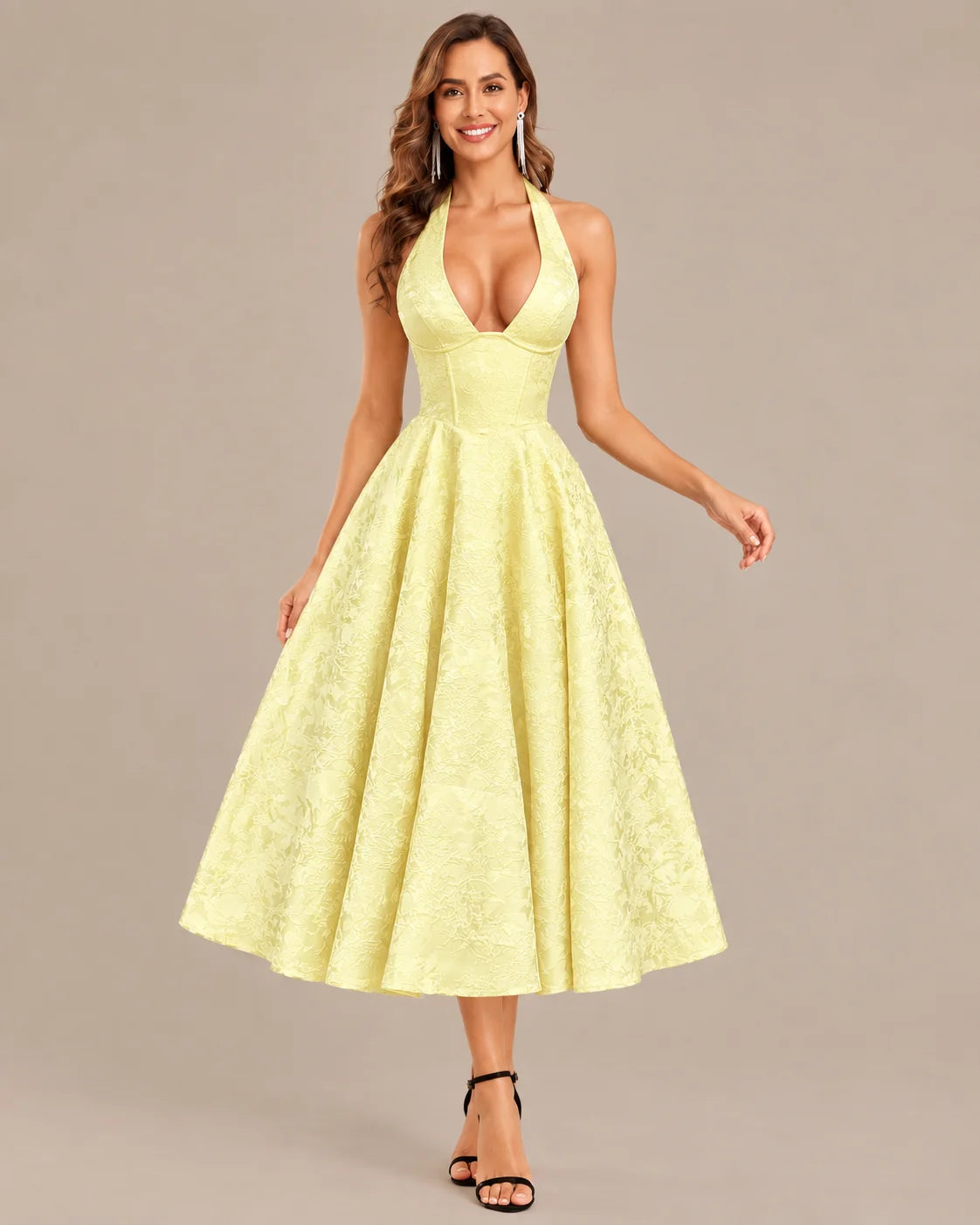 A woman modeling a pale yellow halter midi dress with a deep V-neckline, fitted waist, and full flared skirt, photographed against a plain warm gray studio background.