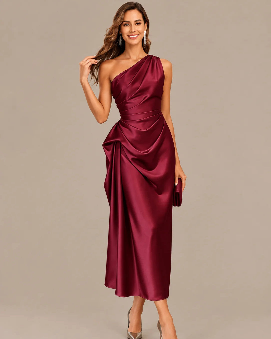 A woman modeling a one-shoulder midi dress in deep burgundy satin, paired with silver pointed-toe heels and holding a matching clutch, set against a solid warm gray studio background.