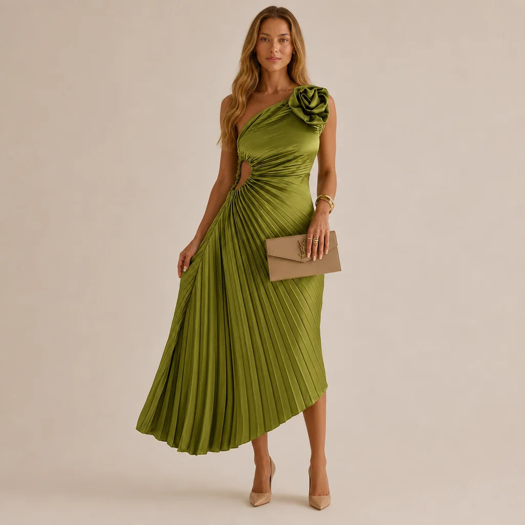 A woman models a one-shoulder olive green midi dress with a pleated skirt and a large fabric flower detail on the sleeve. She is holding a taupe clutch and wearing nude heels, set against a solid beige studio background.