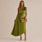 A woman models a one-shoulder olive green midi dress with a pleated skirt and a large fabric flower detail on the sleeve. She is holding a taupe clutch and wearing nude heels, set against a solid beige studio background.