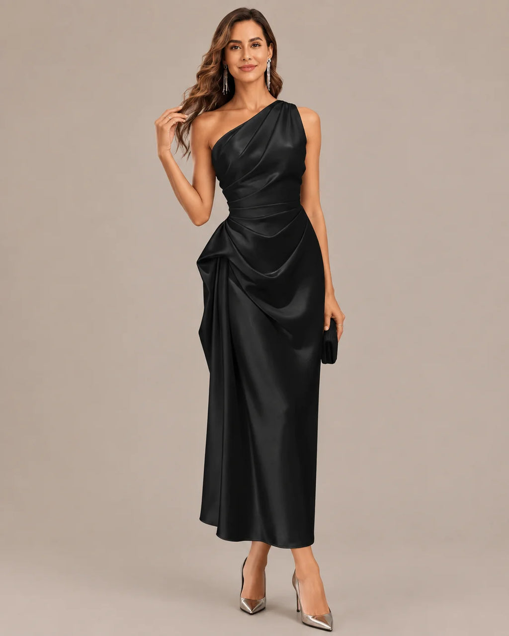 A woman modeling a floor-length black satin one-shoulder gown with a draped, gathered waist and skirt, paired with pointed silver heels against a plain beige studio background.