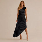 A woman modeling a black one-shoulder midi dress with a pleated skirt and a large black fabric flower detail on the shoulder, photographed against a plain beige studio background.