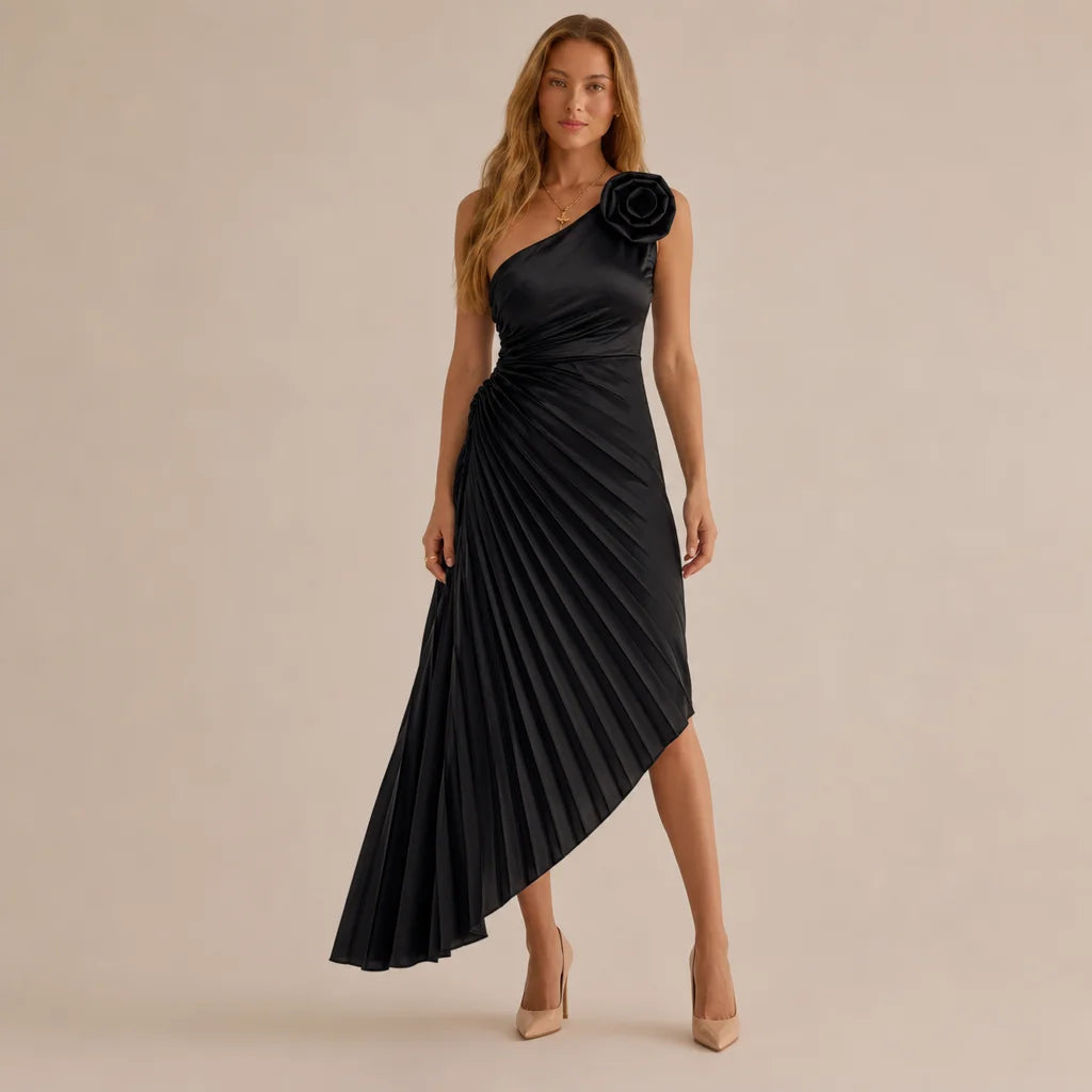 A woman modeling a black one-shoulder midi dress with a pleated skirt and a large black fabric flower detail on the shoulder, photographed against a plain beige studio background.