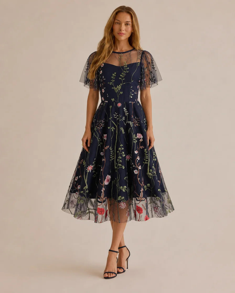 A woman modeling a navy blue midi dress with a floral print, sheer mesh overlay, and short puff sleeves, paired with black strappy high-heeled sandals against a solid beige studio background.