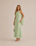 A woman models a sage green, one-shoulder draped maxi dress with a flowing capelet detail on the sleeve, paired with nude strappy high-heeled sandals and a small metallic clutch, set against a solid light beige studio background.