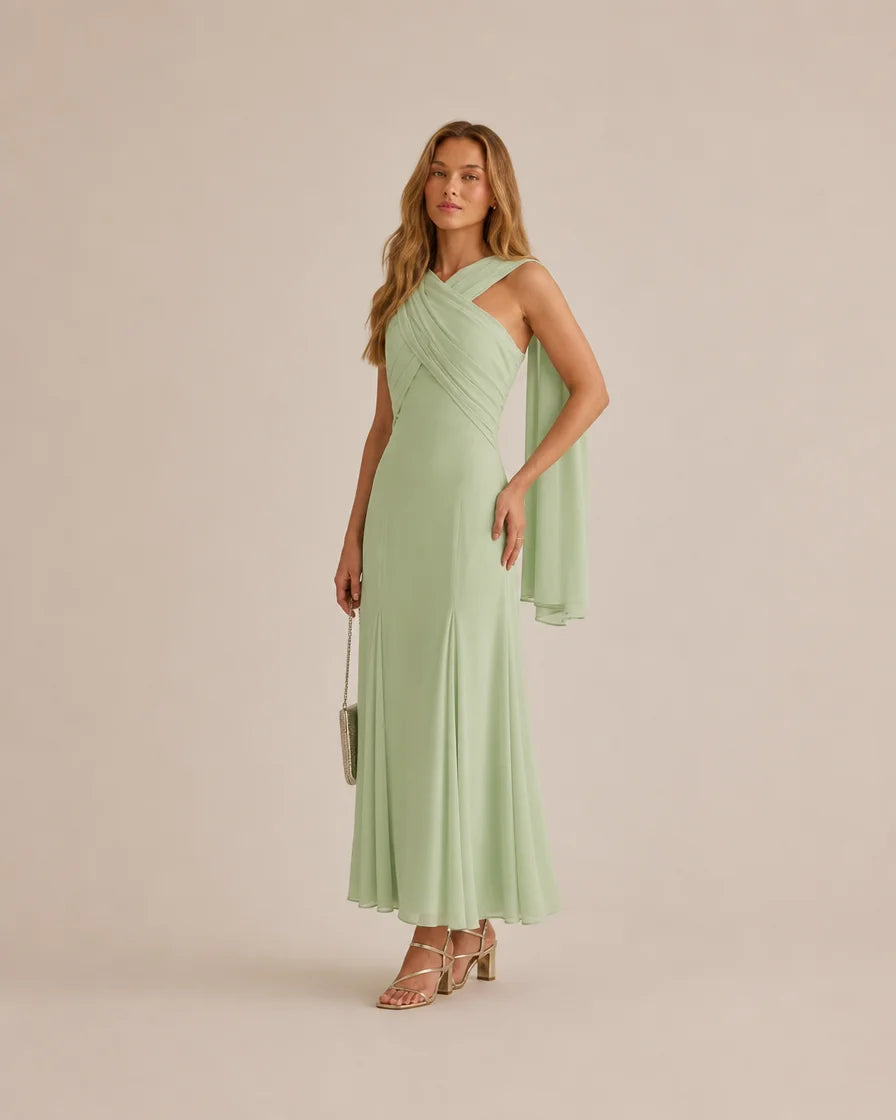 A woman models a sage green, one-shoulder draped maxi dress with a flowing capelet detail on the sleeve, paired with nude strappy high-heeled sandals and a small metallic clutch, set against a solid light beige studio background.