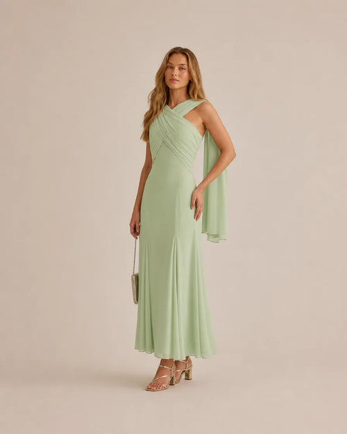 A woman models a sage green, one-shoulder draped maxi dress with a flowing capelet detail on the sleeve, paired with nude strappy high-heeled sandals and a small metallic clutch, set against a solid light beige studio background.