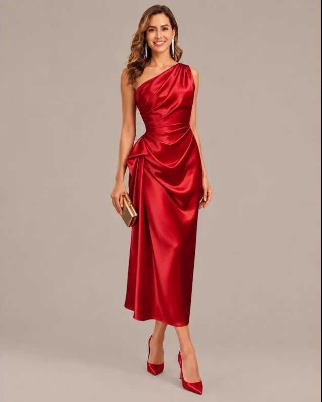 A woman modeling a red satin one-shoulder midi dress with a draped, gathered bodice and skirt, styled with red pointed-toe heels and a small gold clutch against a neutral gray studio background.
