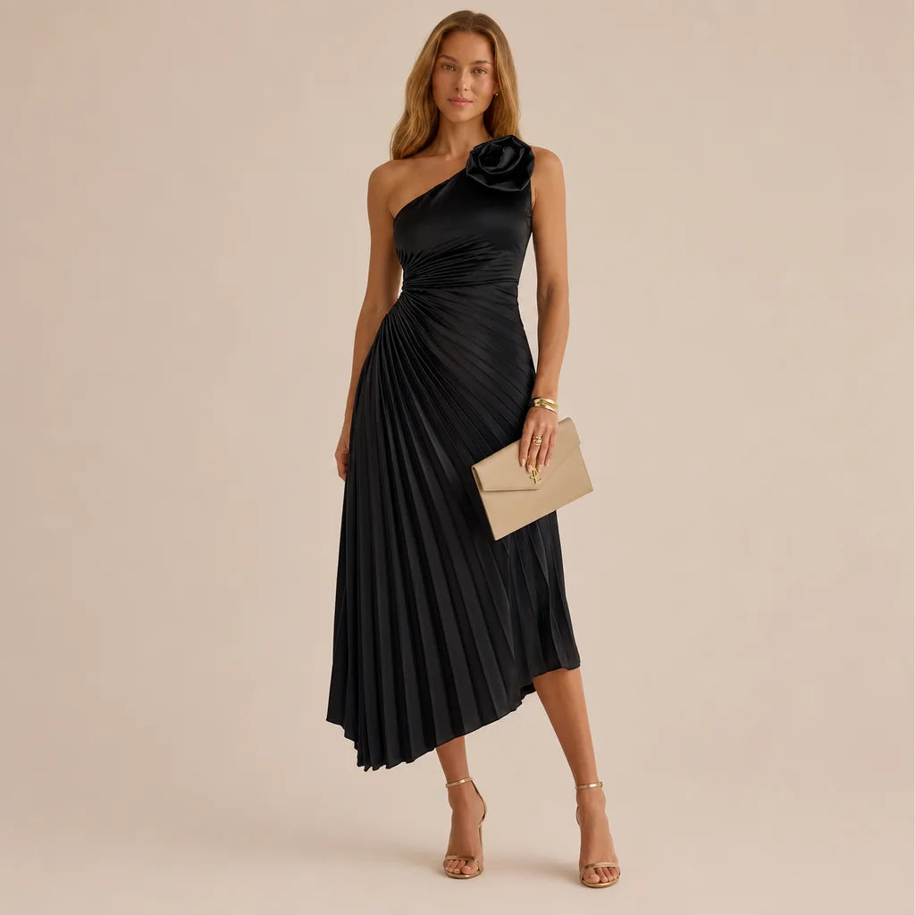 A woman models a black one-shoulder midi dress with a pleated skirt and a large fabric flower detail on the shoulder, paired with nude high heels and a beige clutch, against a solid beige studio background.