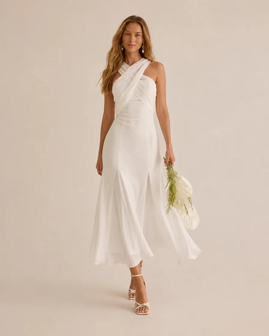 A woman models a white one-shoulder midi dress with a draped neckline and flowing skirt, holding a small white floral bouquet against a solid light beige studio background.