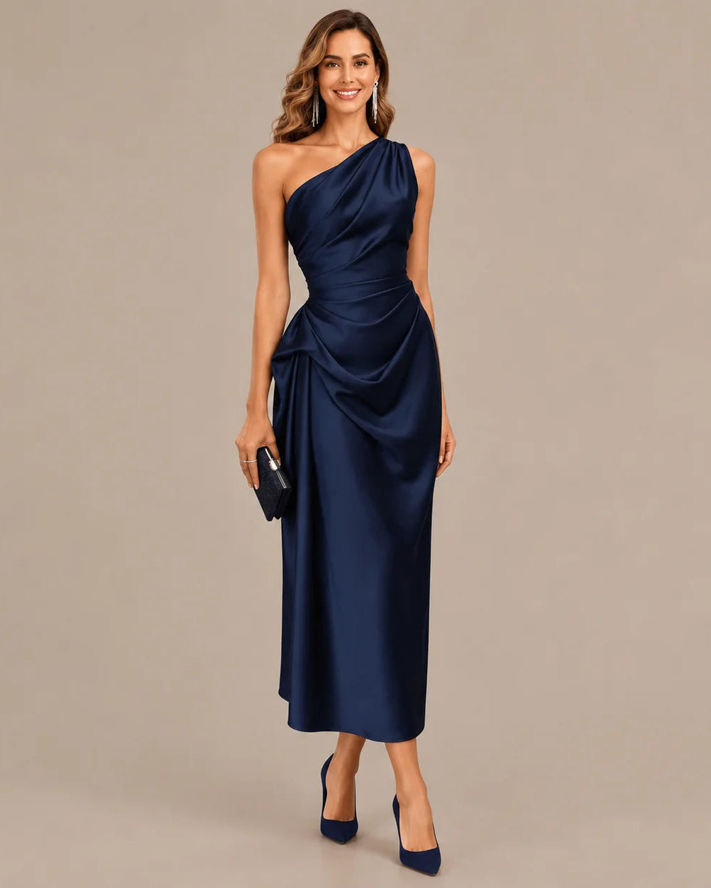 A woman modeling a navy blue one-shoulder midi dress with a draped satin-like finish, paired with navy heels and a small black clutch, against a solid beige studio background.