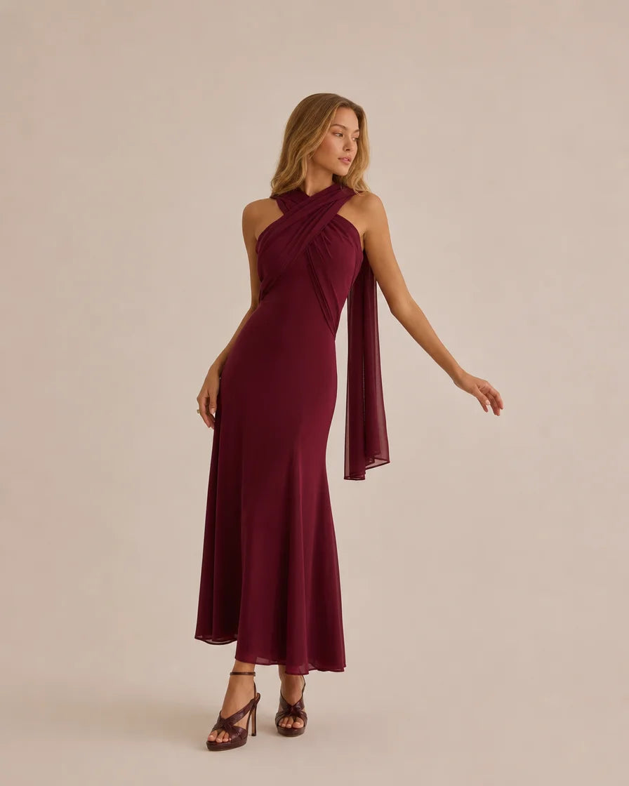 A woman modeling a sleeveless, floor-length halter gown in deep burgundy with a draped chiffon shoulder overlay, set against a solid light beige studio background.