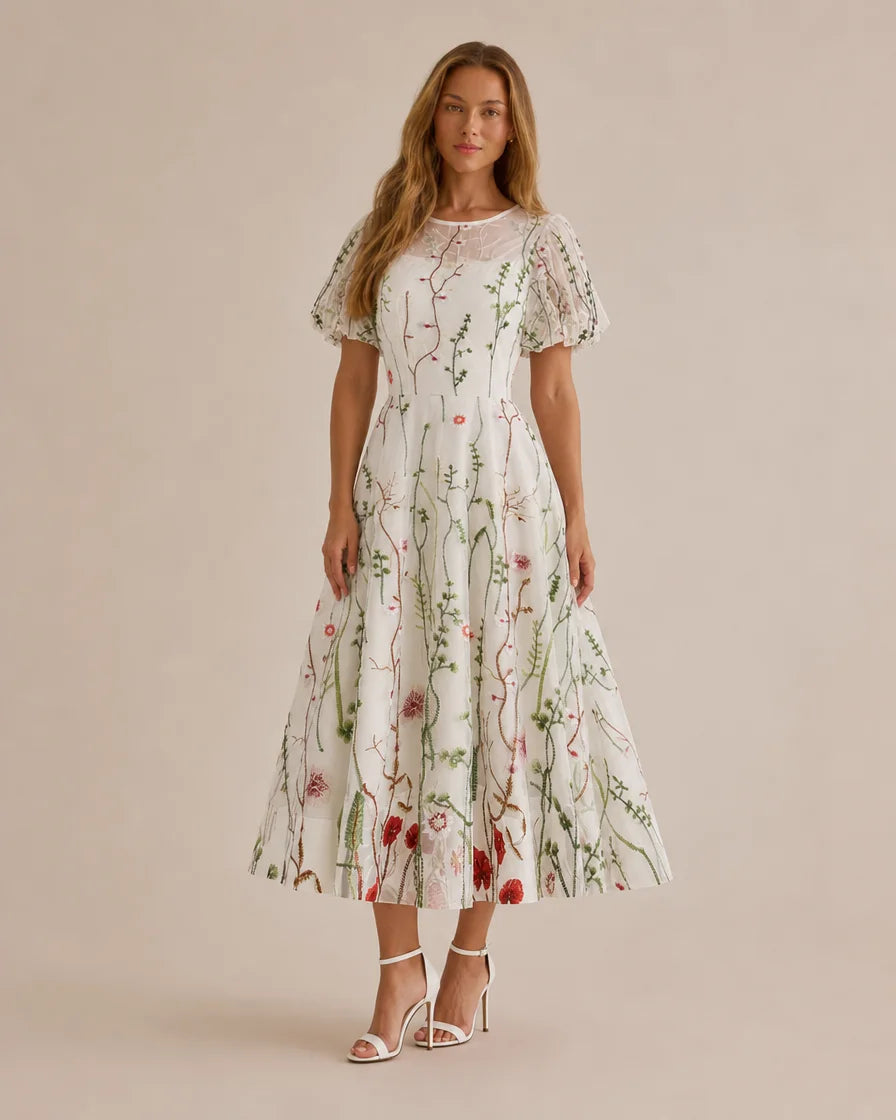A woman modeling a white midi dress with a green and red floral vine pattern, featuring short puff sleeves and a sheer lace neckline, set against a solid beige studio background.