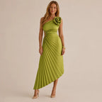 A woman models a one-shoulder midi dress in olive green with a pleated skirt and a large fabric flower detail on the shoulder, set against a plain beige studio background.